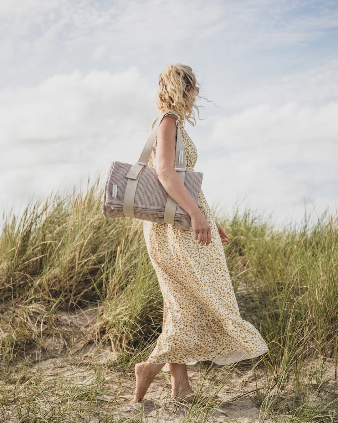 Eine Frau läuft durch die Dünen, über ihrer Schulter hängt am Tragegurt ein zusammengerolltes Strandsofa.