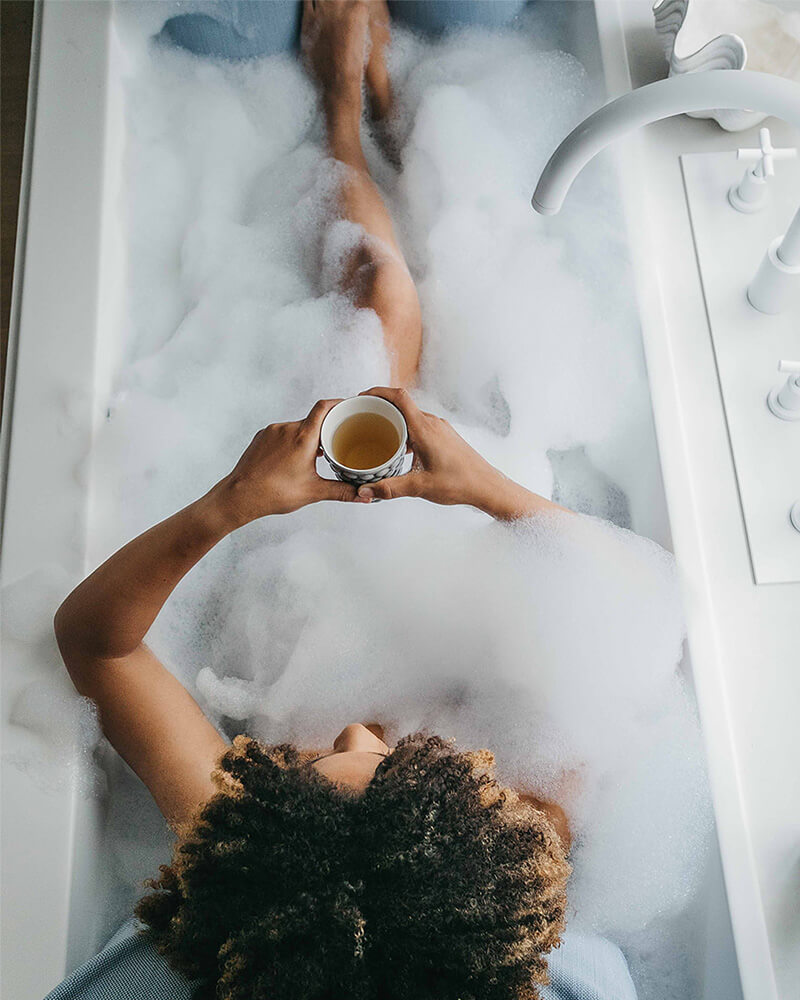 Frau badet mit Rückenkissen (Large) und Fusskissen in luxuriöser freistehender Badewanne. Rücken, Kopf und Füsse ruhen entspannt auf Badesofas (Farbe Blau).