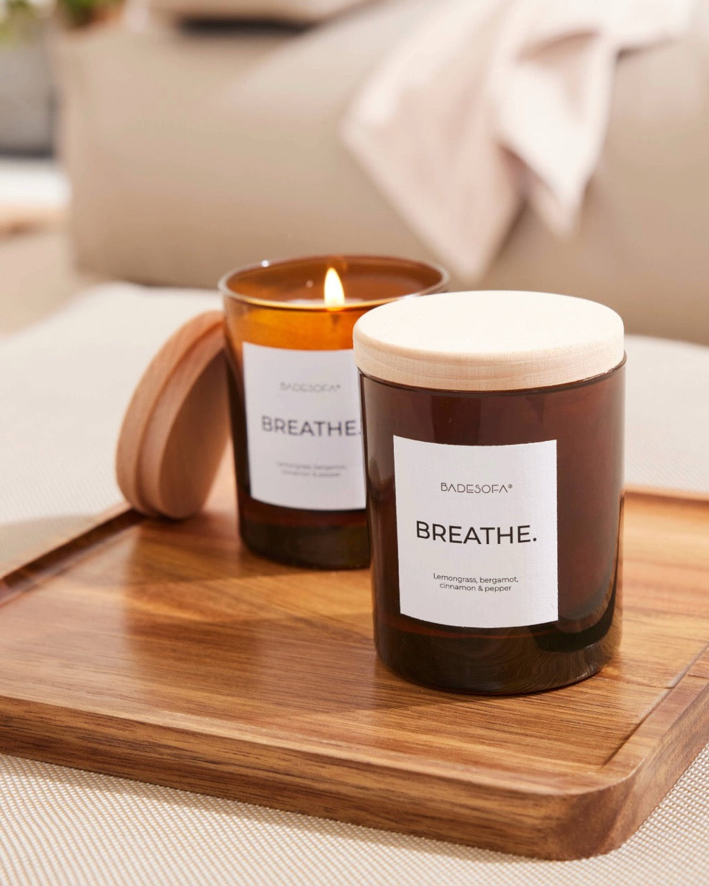 Zwei Duftkerzen von Badesofa (Duftrichtung Breathe) stehen auf einem Holztablett, ein Kerzenglas ist mit einem Holzdecke verschlossen, die andere Kerze ist offen und brennt, der Deckel ist an das Kerzenglas angelehnt.