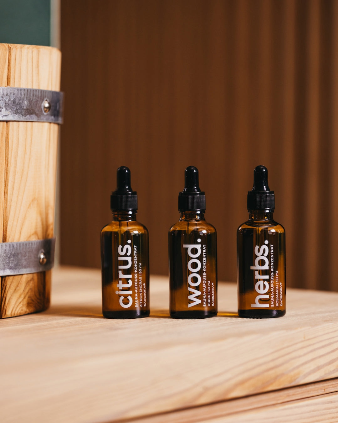 Nahaufnahme von drei kleinen Glasfläschchen mit Pipette, die drei Duftrichtungen Saunaaufguss-Konzentrat enthalten. Die Duftrichtungen sind Wood, Cirtus und Herbs. Neben den Flschen steht ein Holzkübel für Wasser.