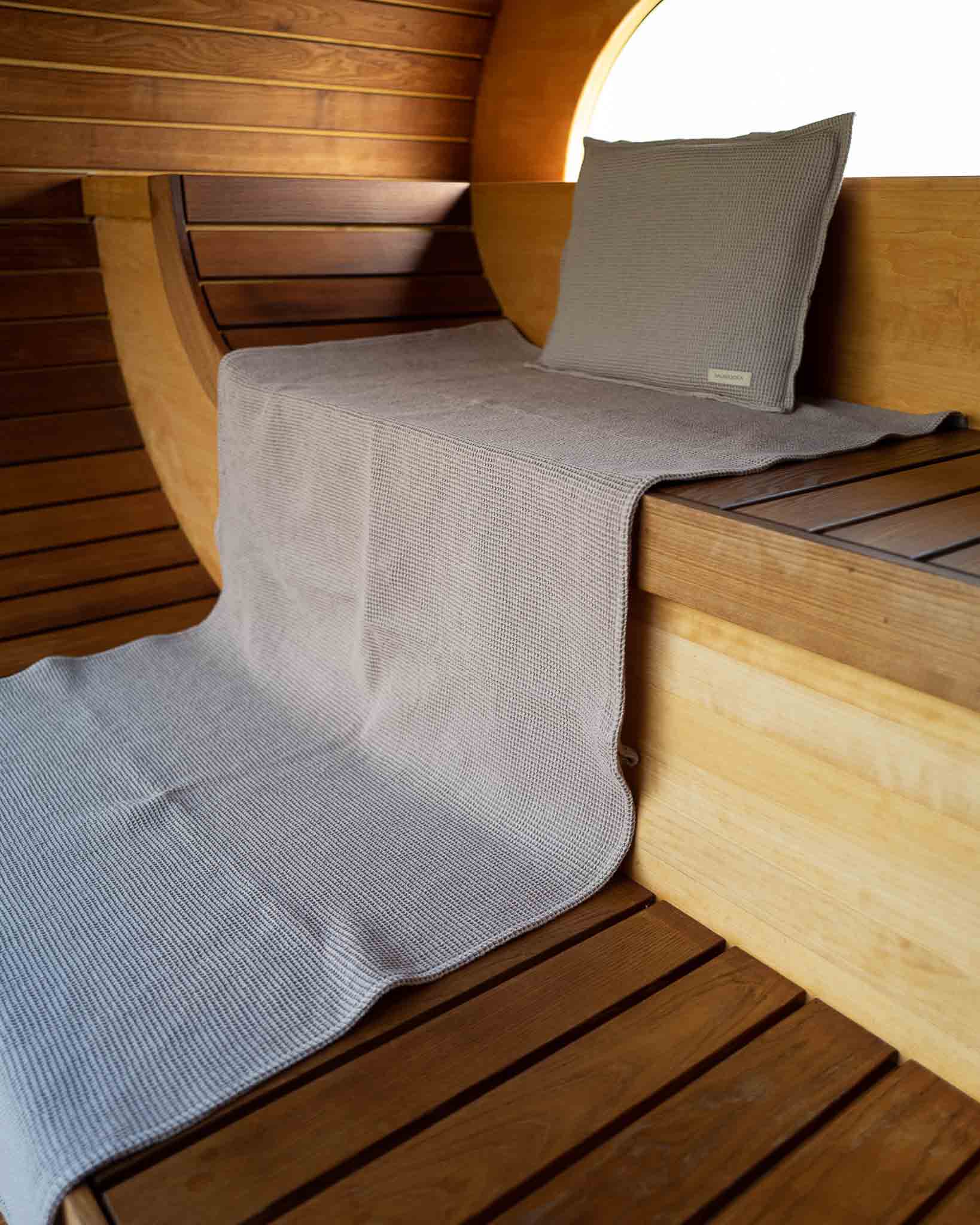 Saunatuch ausgebreitet über eine Saunabank und ein Saunakopfkissen steht aufrecht am Ende der Saunabank in Taupe.