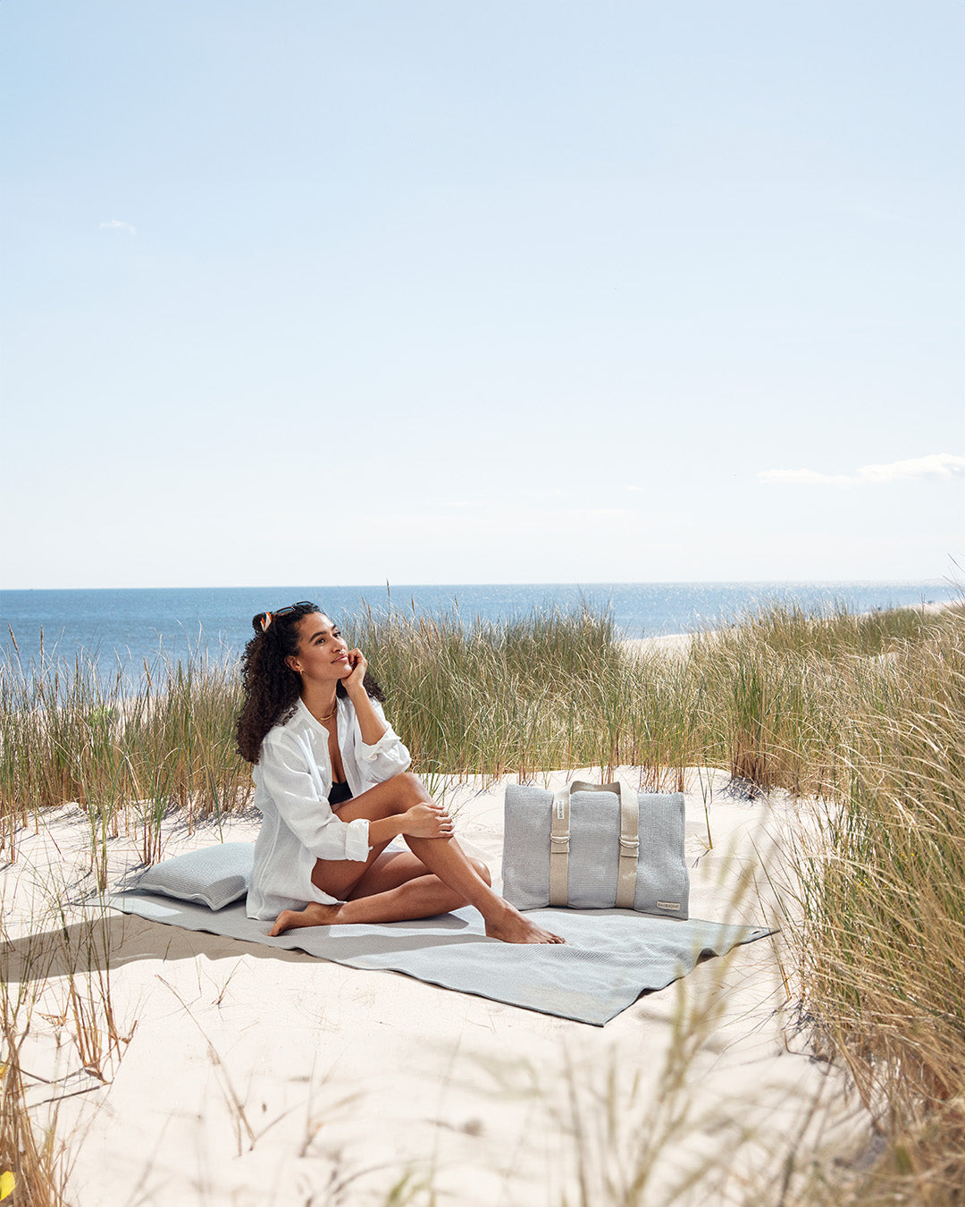 Eine Frau Sitz in den Dünen auf einem Badetuch, hinter ihr liegt das Kopfkissen. Neben ihr steht ein zweites Strandsofa zusammengerollt und gehalten durch den flexiblen Tragegurt.