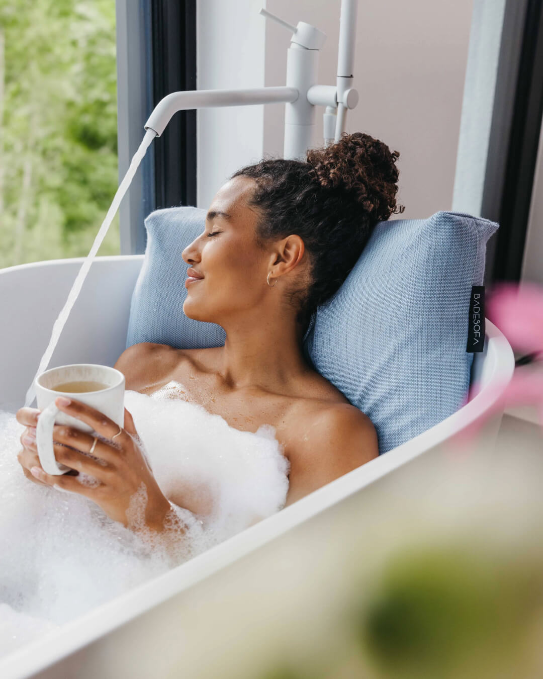 Frau lehnt entspannt an großem Rückenkissen (Large, Blau) in luxuriöser freistehender Badewanne. Das Kissen stützt Kopf, Nacken und Rücken. 