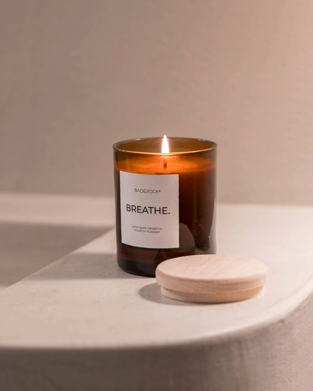 EIne Duftkerzen von Badesofa (Duftrichtung Breathe) steht auf edem Rand eines Betonwaschbeckens. Die Kerze ist offen und brennt, der Holzdeckel liegt vor dem Kerzenglas.