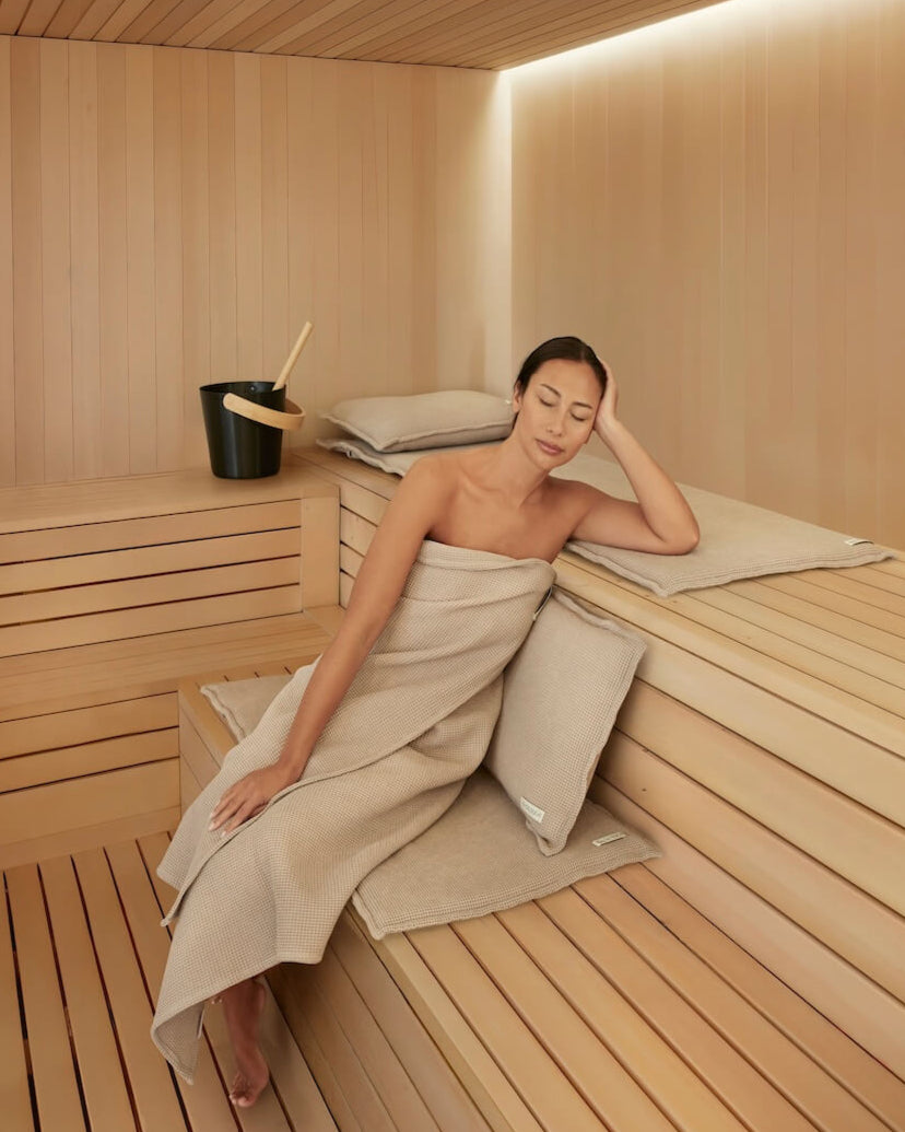 Frau sitz entspannt in der Sauna und leht sich mit dem Rücken an ein Saunasofa Kopfkissen. Sie sitzt auf einer gepolsterten Saunaauflage.