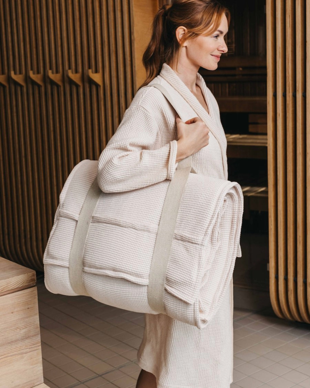 Eine Frau läuft im Bademantel durch einen Wellnessbereich und trägt ein zusammengerolltes Saunasofa Set am Tragegurt über der Schulter. $ Farbe Perlmutt