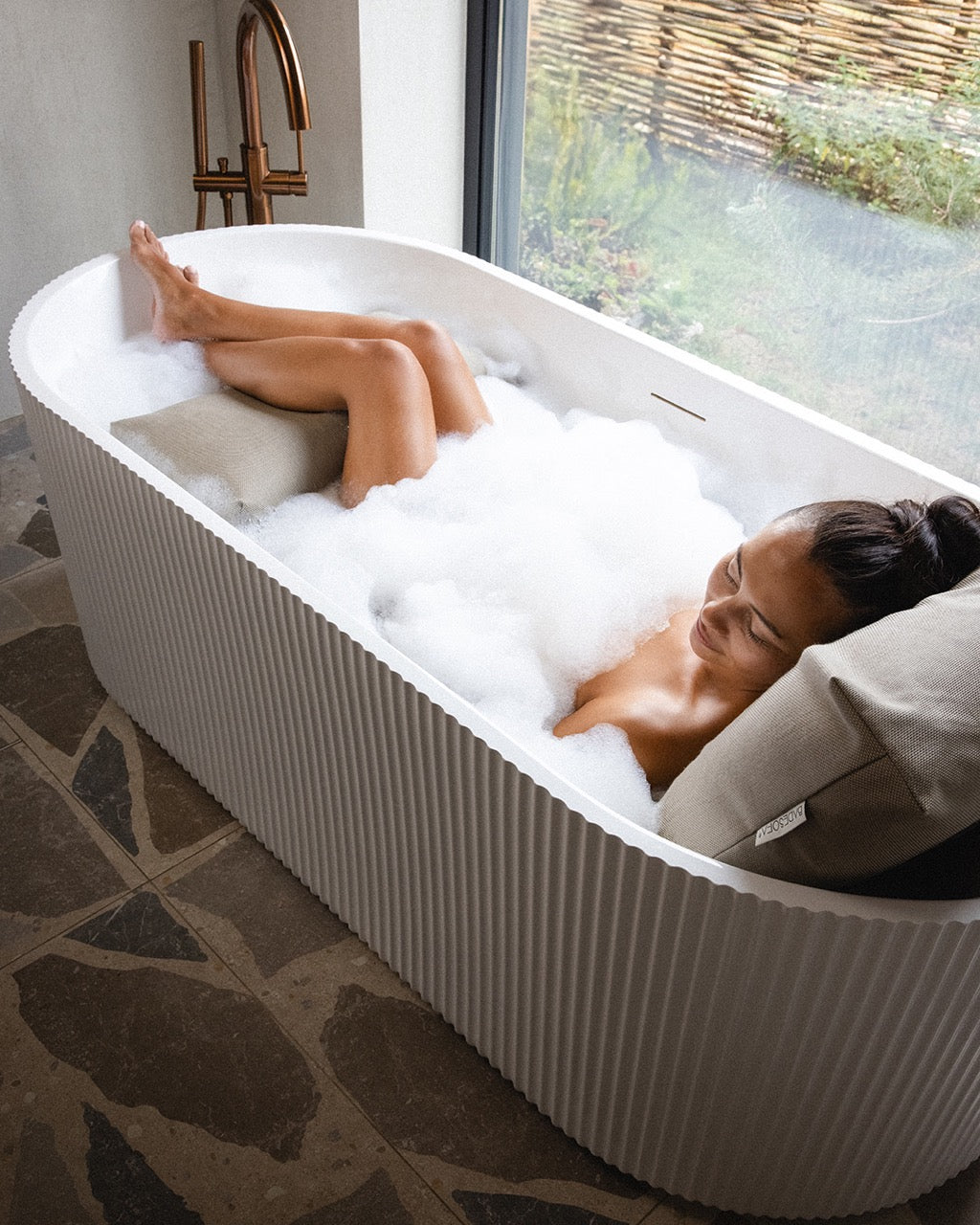 Frau badet mit Rückenkissen (Large) und Fusskissen in luxuriöser freistehender Badewanne. Rücken, Kopf und Füsse ruhen entspannt auf einem Badesofa.