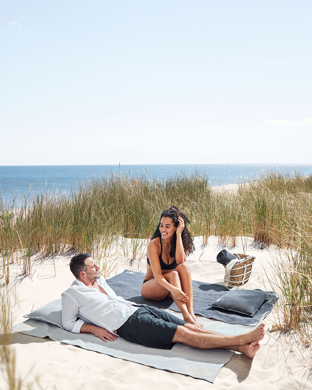 Ein Mann und eine Frau sitzen in den Dünen am Strand, jeder auf einem großen Badetuch. Auf beiden Strandtüchern liegt ein Kopfkissen.