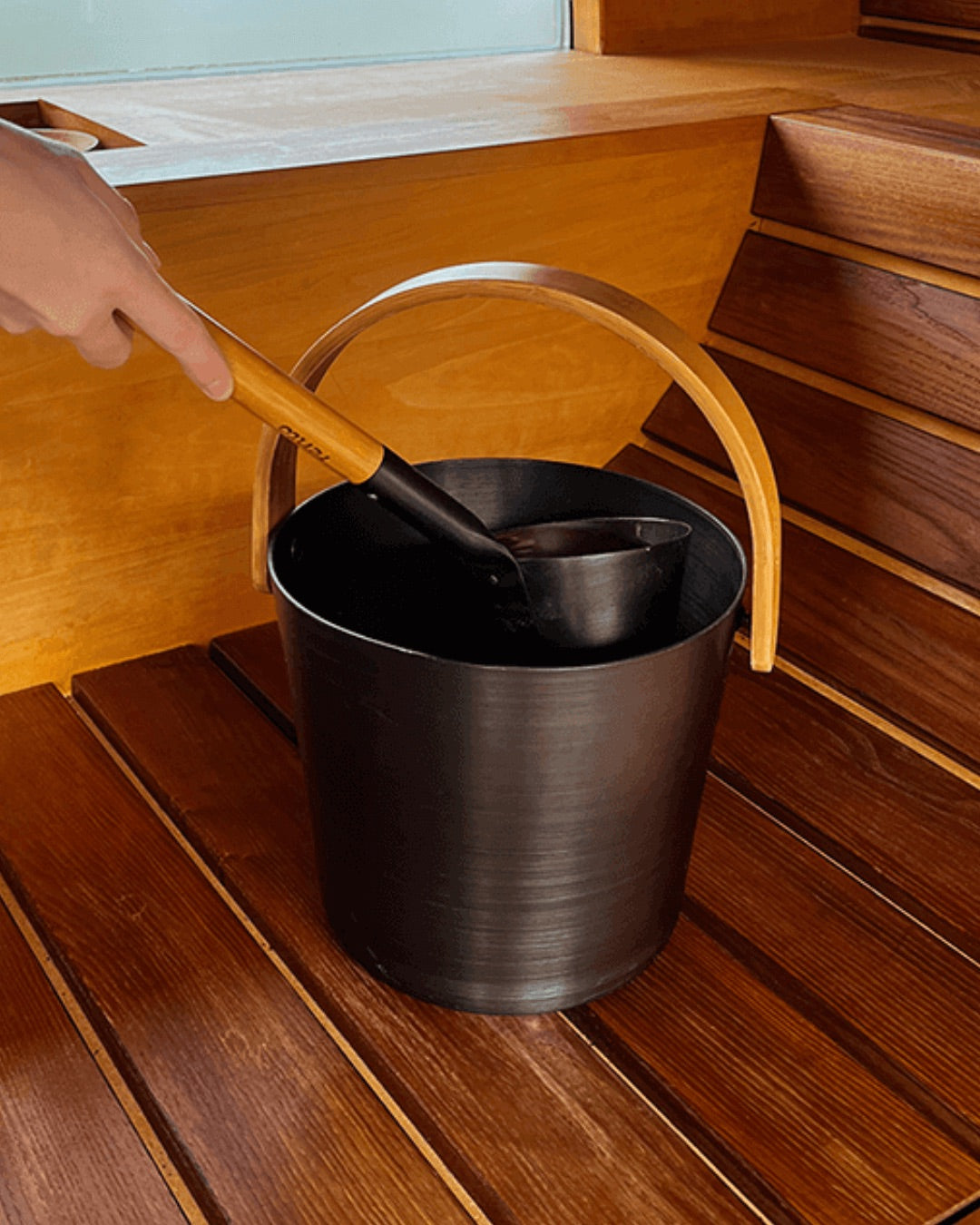 Eine Person schöpft mit der Kelle Wasser aus dem Saunakübel in der Farbe Braun.